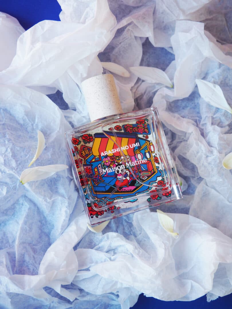 Der Parfumflakon Maison Matine Arashi No Umi aus der Voyager Collection liegt auf hellem Papier mit Blütenblättern. Die farbenfrohe Illustration auf dem Glas zeigt eine verspielte, japanisch inspirierte Szenerie und verweist auf die Sehnsucht nach fernen Horizonten und geheimnisvollen Inseln.