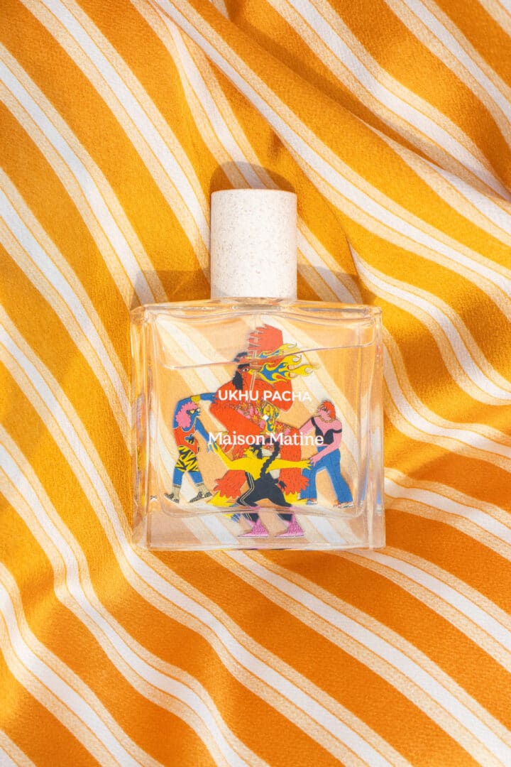 Der Parfumflakon Maison Matine Ukhu Pacha aus der Voyager Collection liegt auf gelb-weiß gestreiftem Stoff. Die Illustration von Caroline Gibaut zeigt farbenfrohe Figuren in Bewegung und symbolisiert Spiritualität, Tanz und Energie, die das Thema des Duftes visuell unterstreichen.