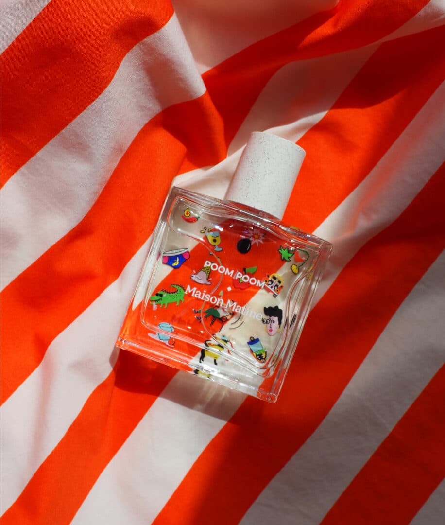 The Maison Matine Poom Poom perfume bottle from the Refresh Collection lies on an orange and white striped fabric. The colorful illustration on the glass shows playful symbols and figures and reflects the energy, joy and lightness of the fragrance.