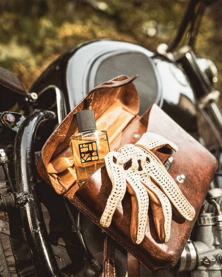 Estora's perfume bottle in a brown leather bag next to cream-colored leather gloves on a motorcycle - a symbol of adventure and style
