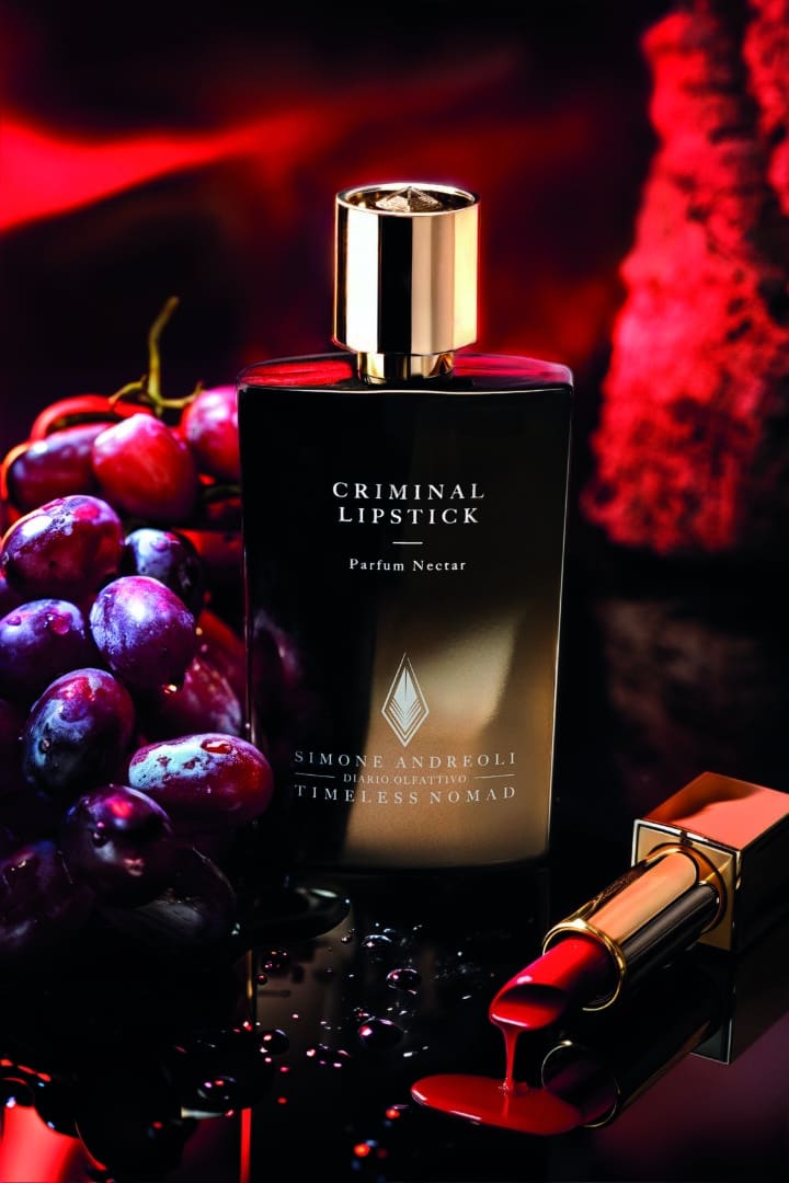Flacon of Criminal Lipstick from the Timeless Nomad Collection by Simone Andreoli, staged with red grapes and an open lipstick on a shiny background against a deep red background.