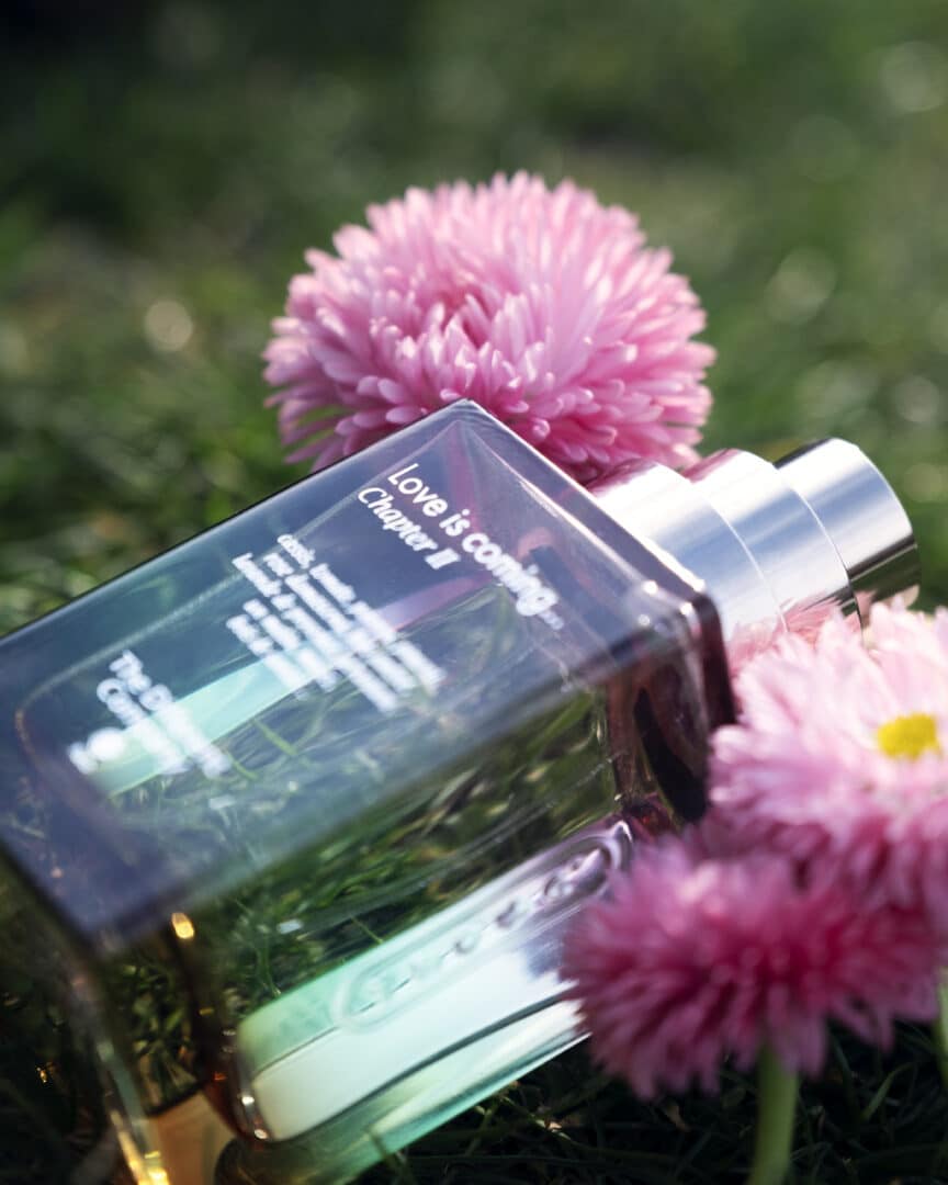 Love is coming … Chapter II von The Different Company – Flakon im Gras mit rosa Blüten, fotografiert im Sonnenlicht