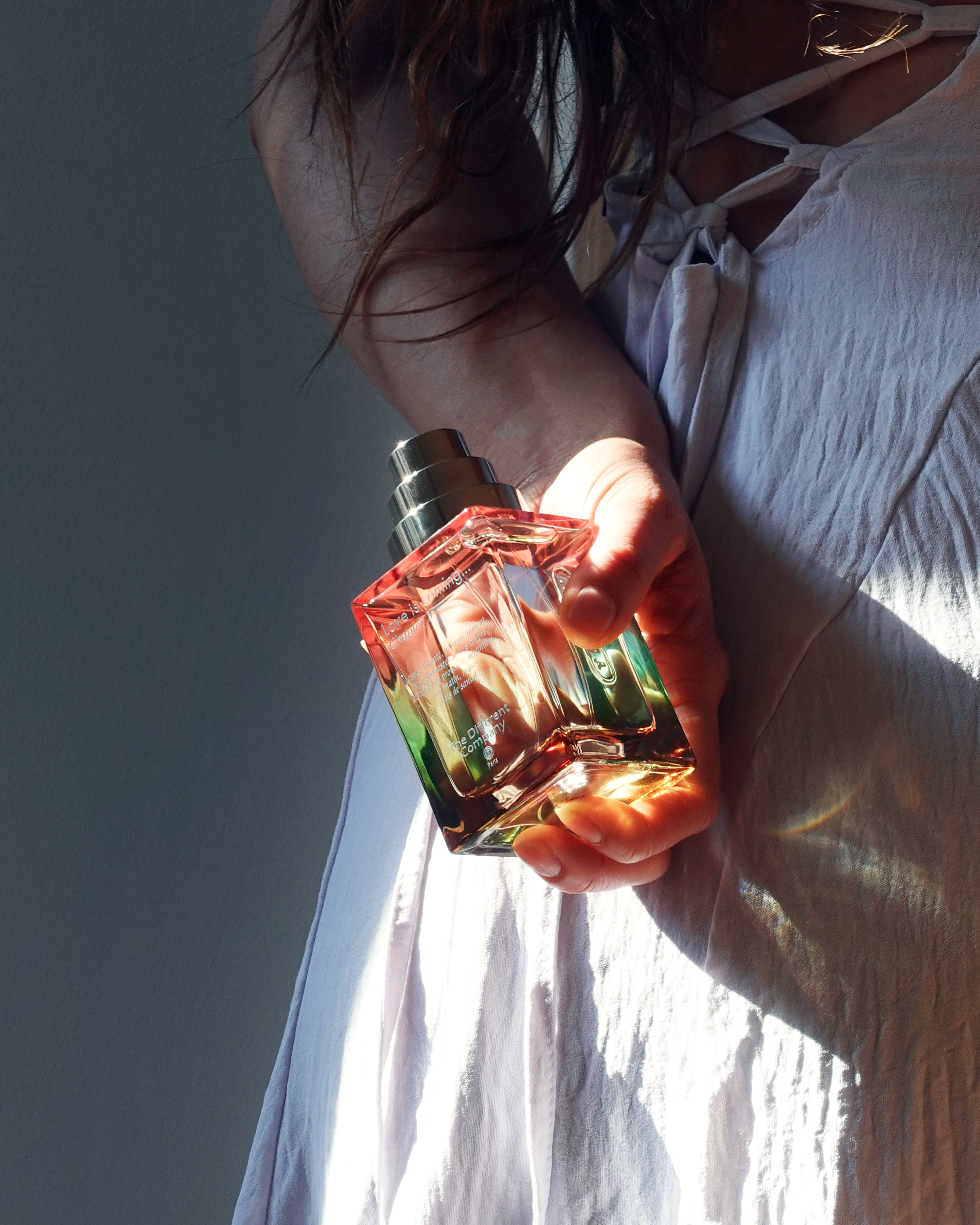 Love is coming … Chapter II von The Different Company – Flakon in der Hand gehalten, sanftes Sonnenlicht auf Kleid und Haut