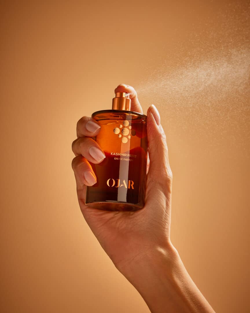 Hand sprüht den Duft Kashmir Print Eau de Parfum von Ojar – Nahaufnahme mit feinem Sprühnebel vor warmem, sandfarbenem Hintergrund.