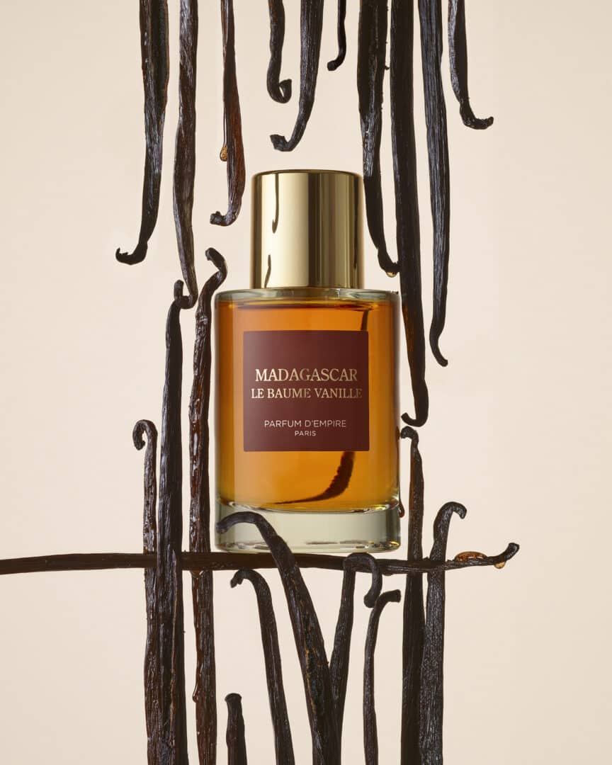 Flakon von Madagascar Le Baume Vanille von Parfums d’Empire, umgeben von dunklen Vanilleschoten – warmer, bernsteinfarbener Duft mit goldener Kappe.