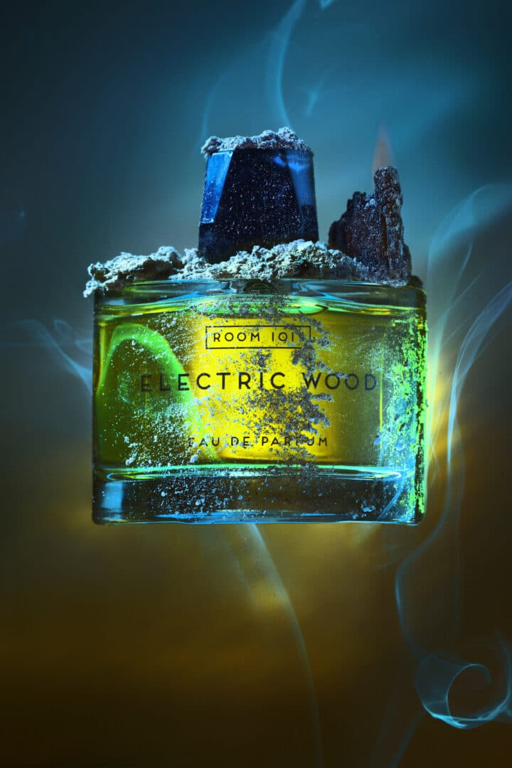Flakon von Room 1015 Electric Wood Eau de Parfum, inszeniert mit Holz- und Ascheelementen vor blau-goldenem Licht – Symbol für Hitze, Rauch und Musikenergie.