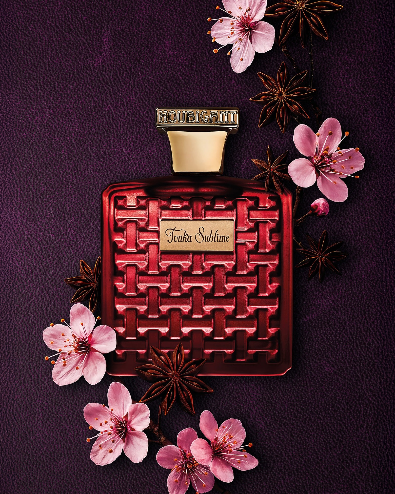 Red bottle of Tonka Sublime by Houbigant on a dark background, surrounded by star anise and pink flowers. Luxurious perfume with notes of tonka bean, almond and amber from the Les Ambres collection.