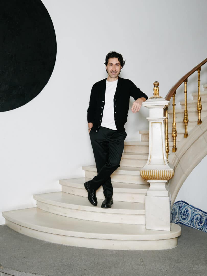 Elad Yifrach steht auf einer weißen Marmortreppe mit goldenen Geländerdetails und lehnt entspannt am Treppenpfosten.