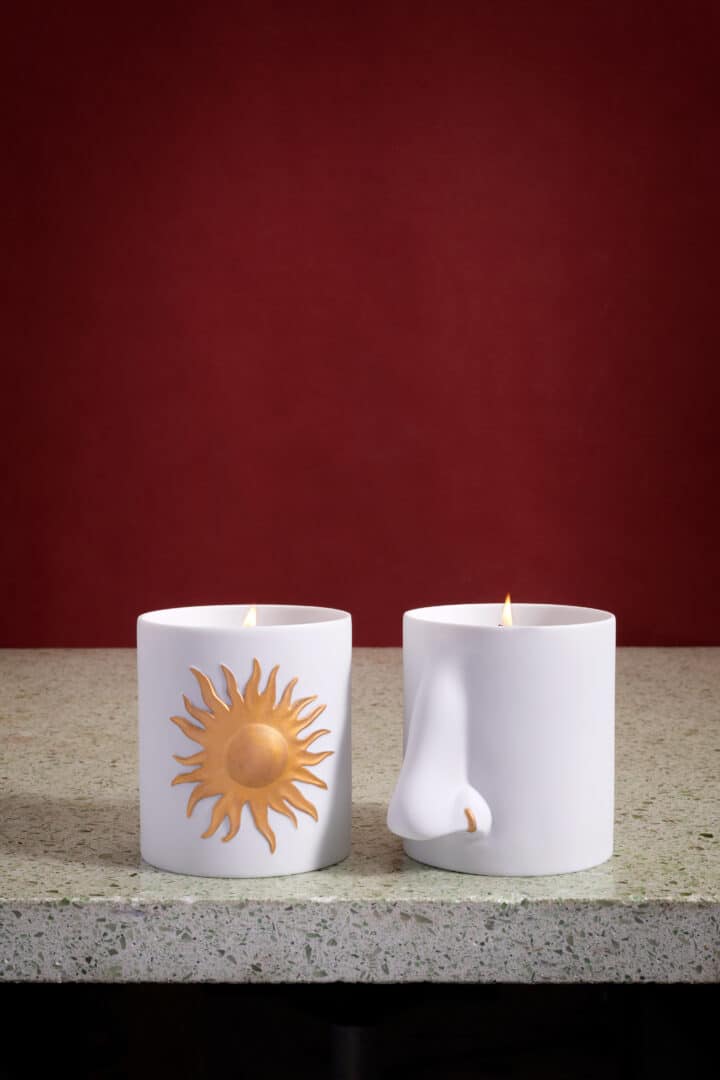 Zwei weiße L’Objet Duftkerzen aus Porzellan auf einer Steinoberfläche vor bordeauxrotem Hintergrund, eine mit goldener Sonne, eine mit skulpturaler Nasendetailierung.