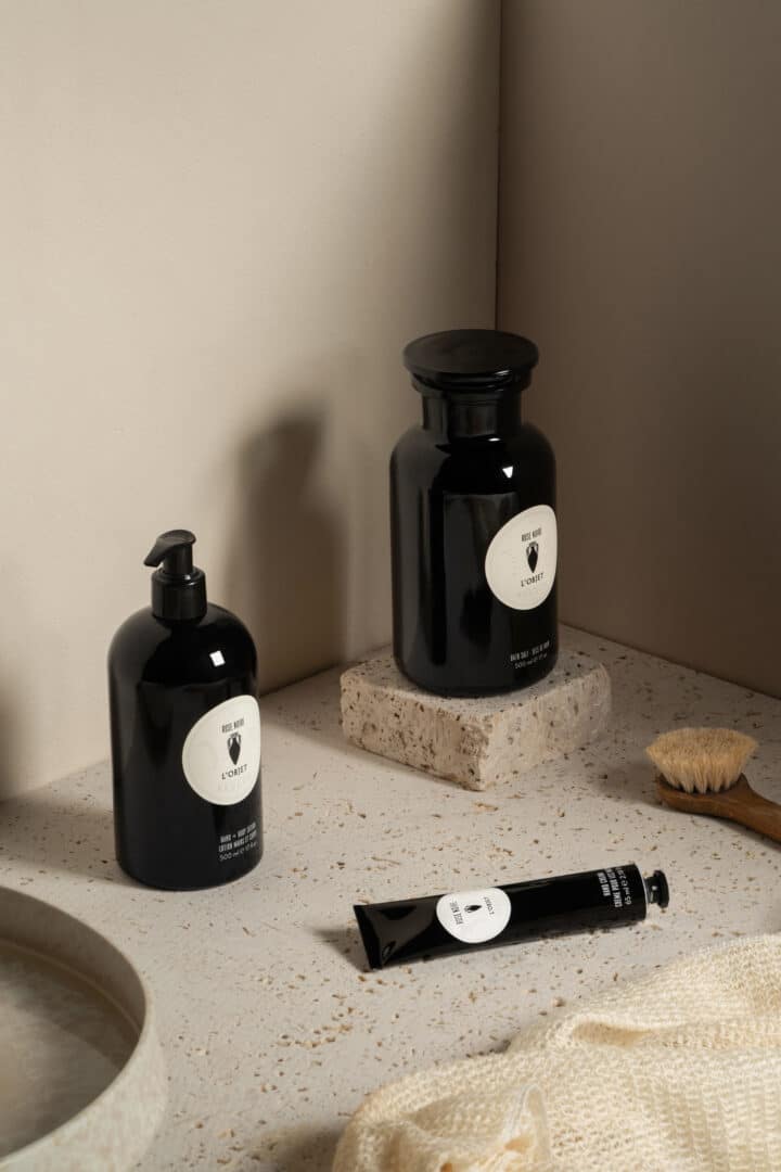 L’Objet Hand- und Körperpflegeprodukte in schwarzen Flakons auf einer natursteinfarbenen Ablage, arrangiert mit Bürste und Texturen in einem minimalistischen Badezimmer-Setting.