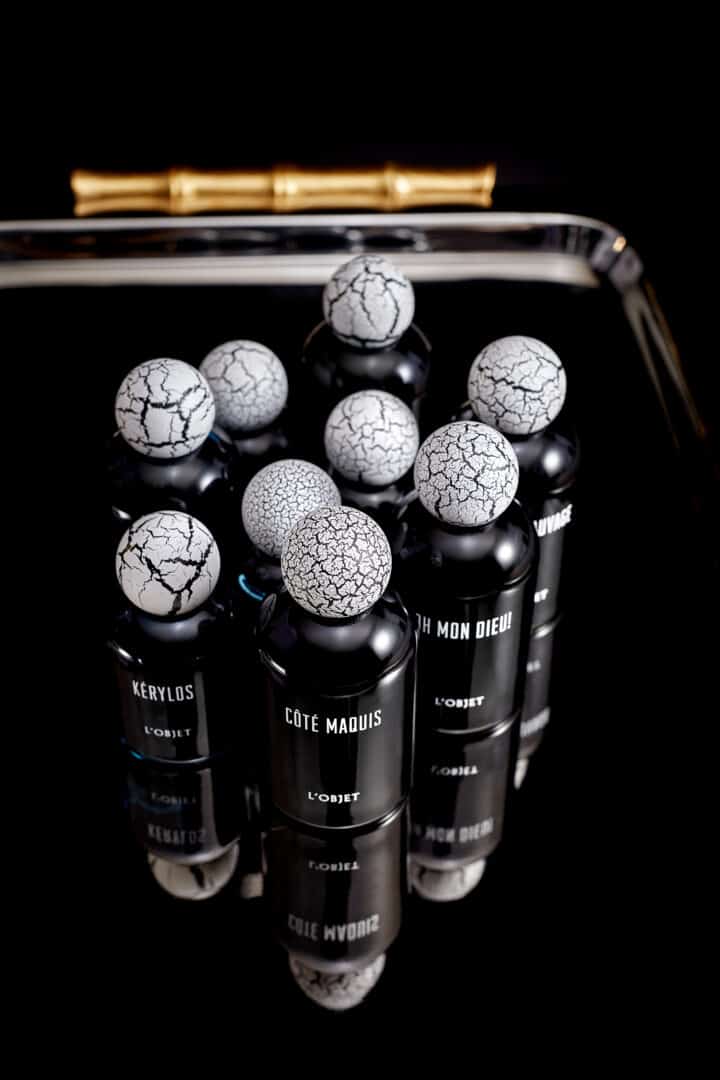 Mehrere schwarze L’Objet Parfumflakons mit weißen, rissigen Kugelkappen auf schwarzer spiegelnder Oberfläche, arrangiert in einer dichten Gruppe mit goldenem Objekt im Hintergrund.