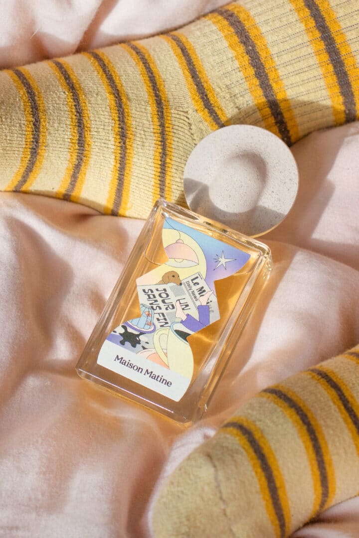 Maison Matine Parfumflakon auf weicher Bettwäsche, umgeben von gelb gestreiften Socken