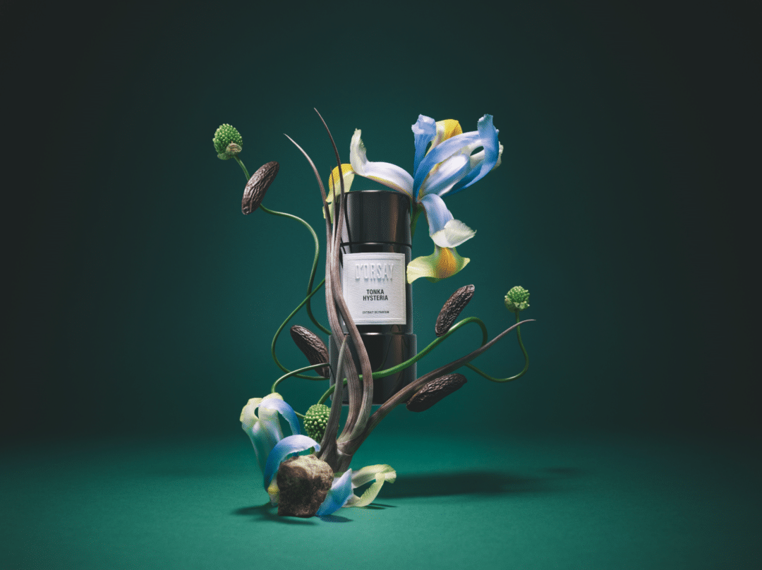 Flakon von ORSAY Tonka Hysteria Extrait de Parfum, inszeniert mit Irisblüten, Tonkabohnen und botanischen Elementen vor grünem Hintergrund