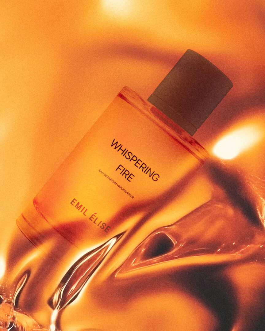 Whispering Fire by Emil Élise - a luminous, warm fragrance in a flowing light setting Whispering Fire by Emil Élise in a transparent glass flacon, surrounded by flowing, golden-orange light and warmth effects