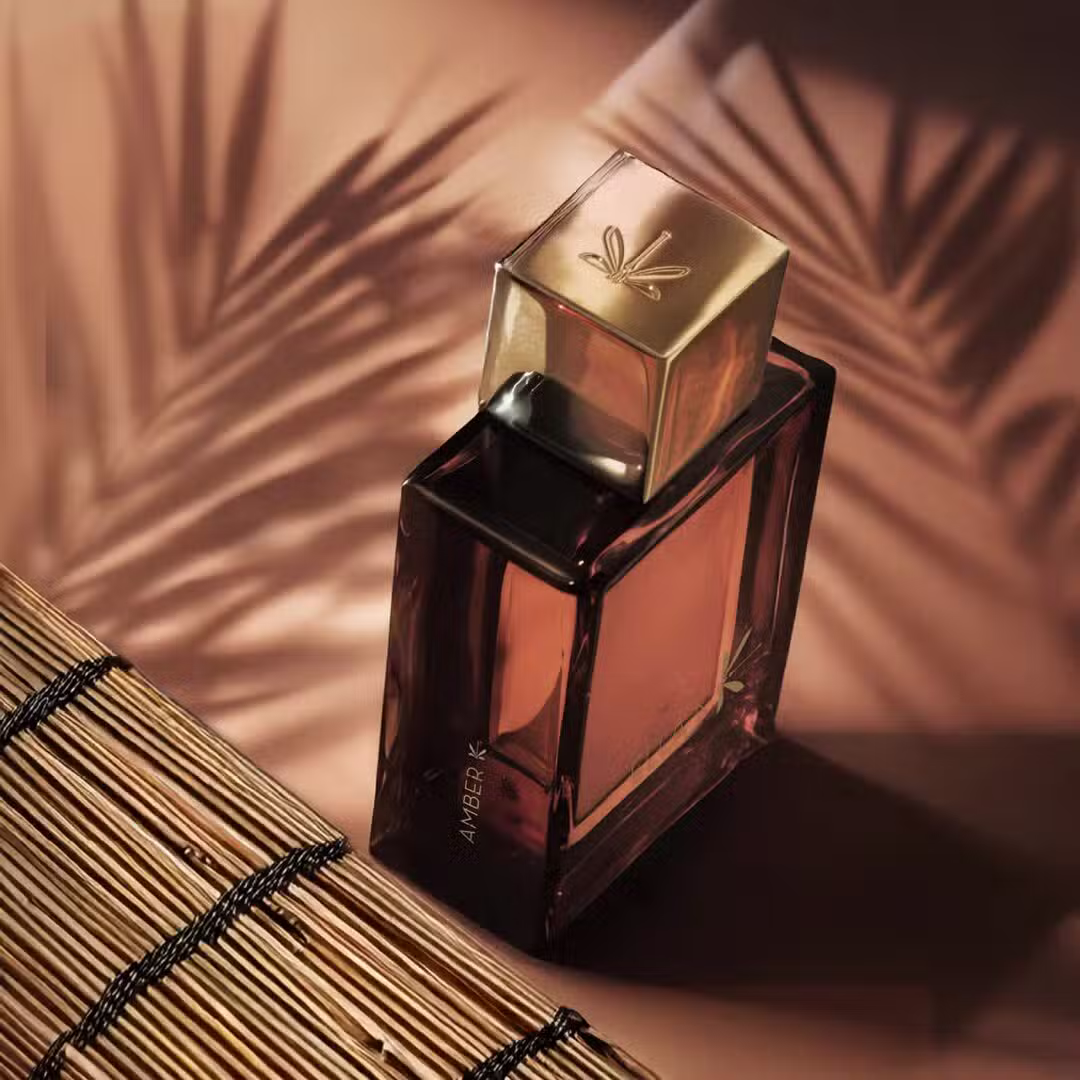 Amber K von Ella K – warmer, sinnlicher Duft mit orientalischer Anmutung Amber K von Ella K – Parfumflakon mit warm schimmerndem Amberton, inszeniert mit Licht- und Palmblatt-Schatten auf natürlichem Untergrund
