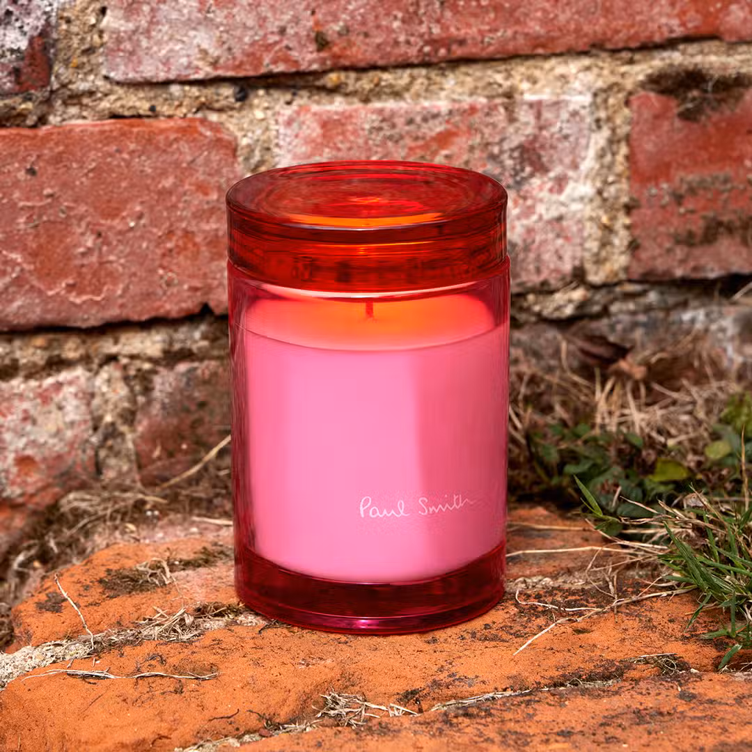 Paul Smith Duftkerze – moderne Farbe trifft auf entspannte Home-Fragrance-Ästhetik Rosafarbene Duftkerze von Paul Smith im transparenten roten Glas vor rustikaler Backsteinwand im Außenbereich