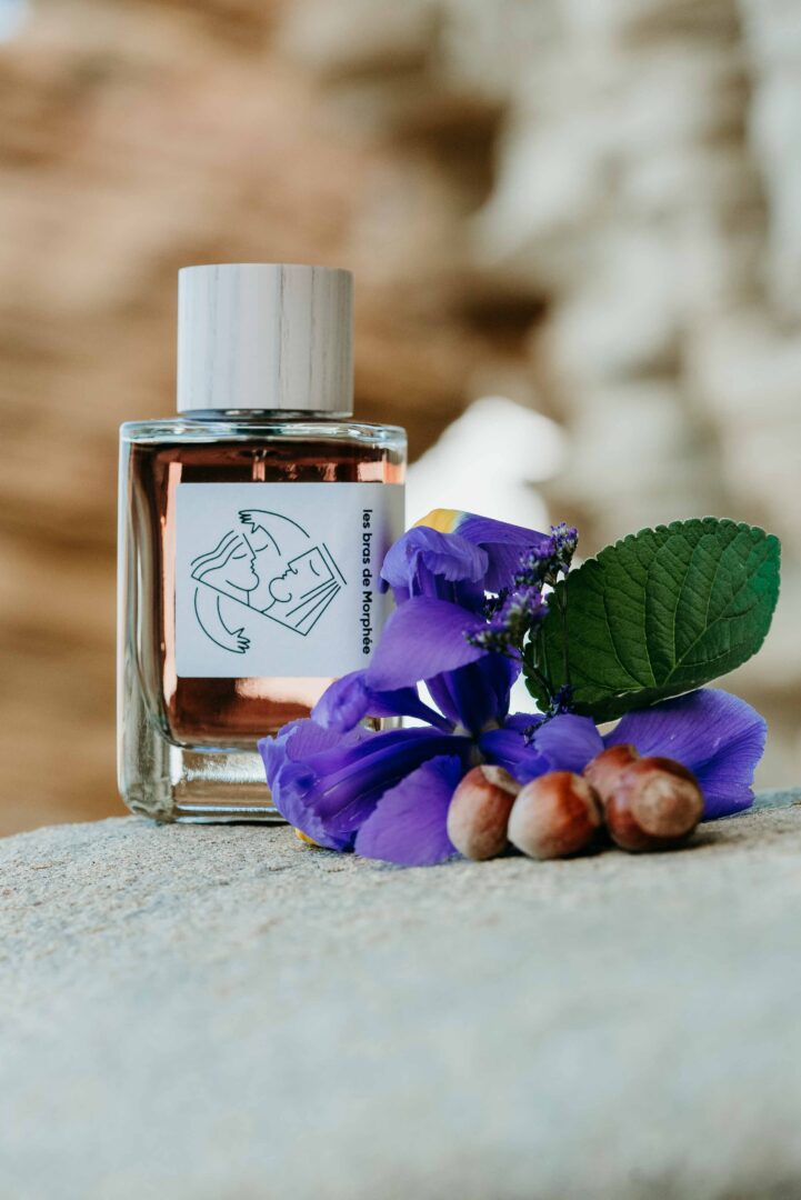 A bottle of Hellenist Paris’s “Les Bras de Morphée” on a stone surface, surrounded by purple iris blossoms, green leaves, and hazelnuts. The presentation highlights the composition’s key fragrance notes and underscores its elegant aesthetic, inspired by Greek mythology.
