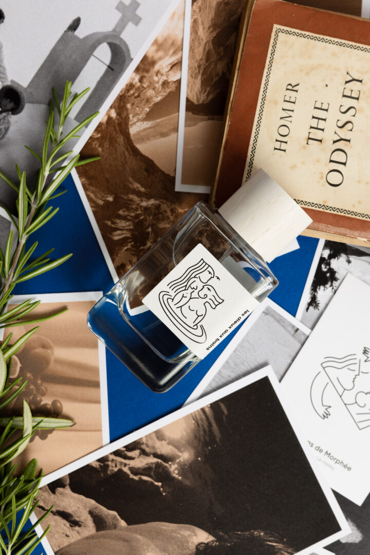 A bottle of Hellenist Paris’s “Les Dieux aux Bains” set against photographs, a copy of Homer’s *Odyssey*, and Mediterranean rosemary sprigs. The display evokes the world of the fragrance, inspired by Greek mythology and culture.