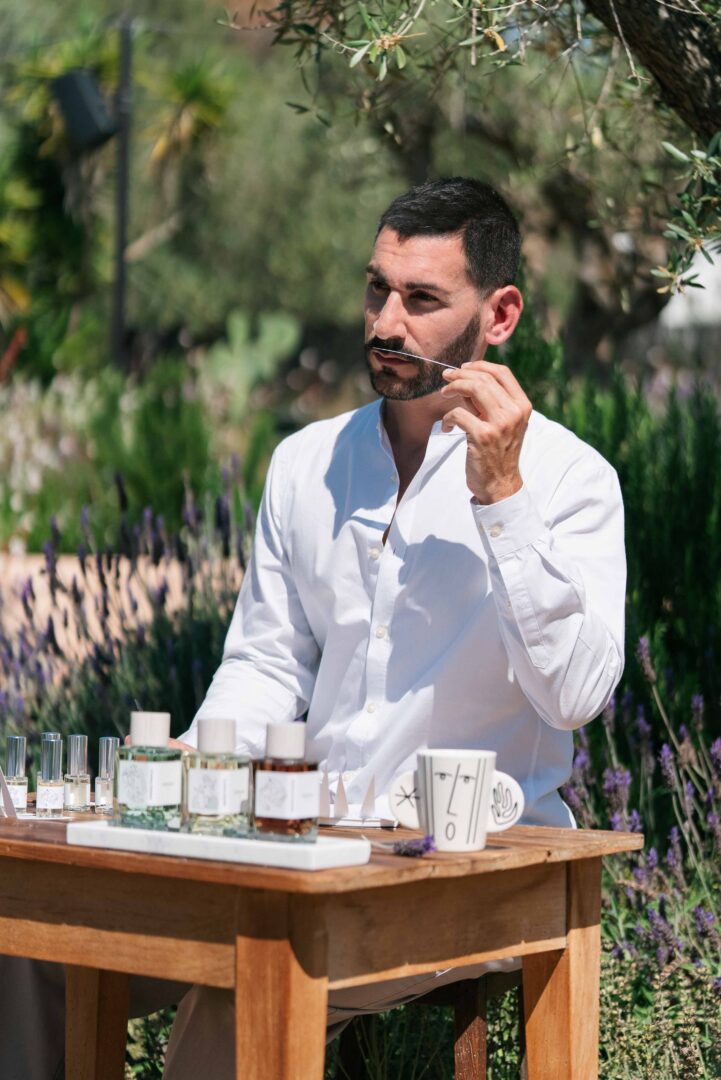 Jean-David Jacoby, founder of Hellenist Paris, sits at a wooden table outdoors, examining a scent strip. In front of him are several perfume bottles, surrounded by Mediterranean vegetation and blooming lavender.