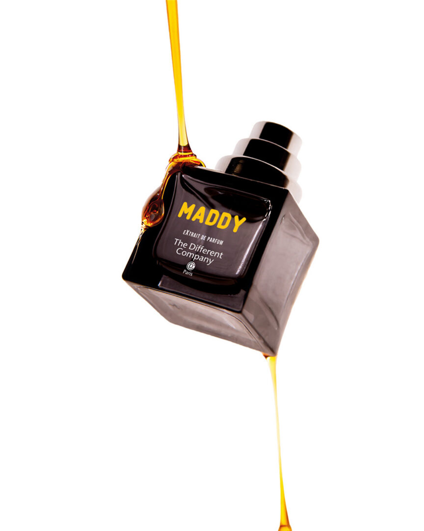 Maddy von The Different Company als Extrait de Parfum. Schwarzer Glasflakon der Extraits Extrêmes Collection, schräg vor hellem Hintergrund arrangiert und von goldfarbenem Honig umflossen. Das Motiv unterstreicht den gourmandigen Charakter des Duftes.