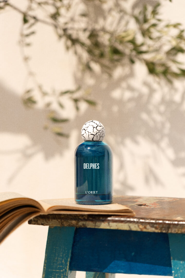 "Delphes" by L'Objet, depicted as a bottle resting on an open book against a backdrop of Mediterranean olive branches