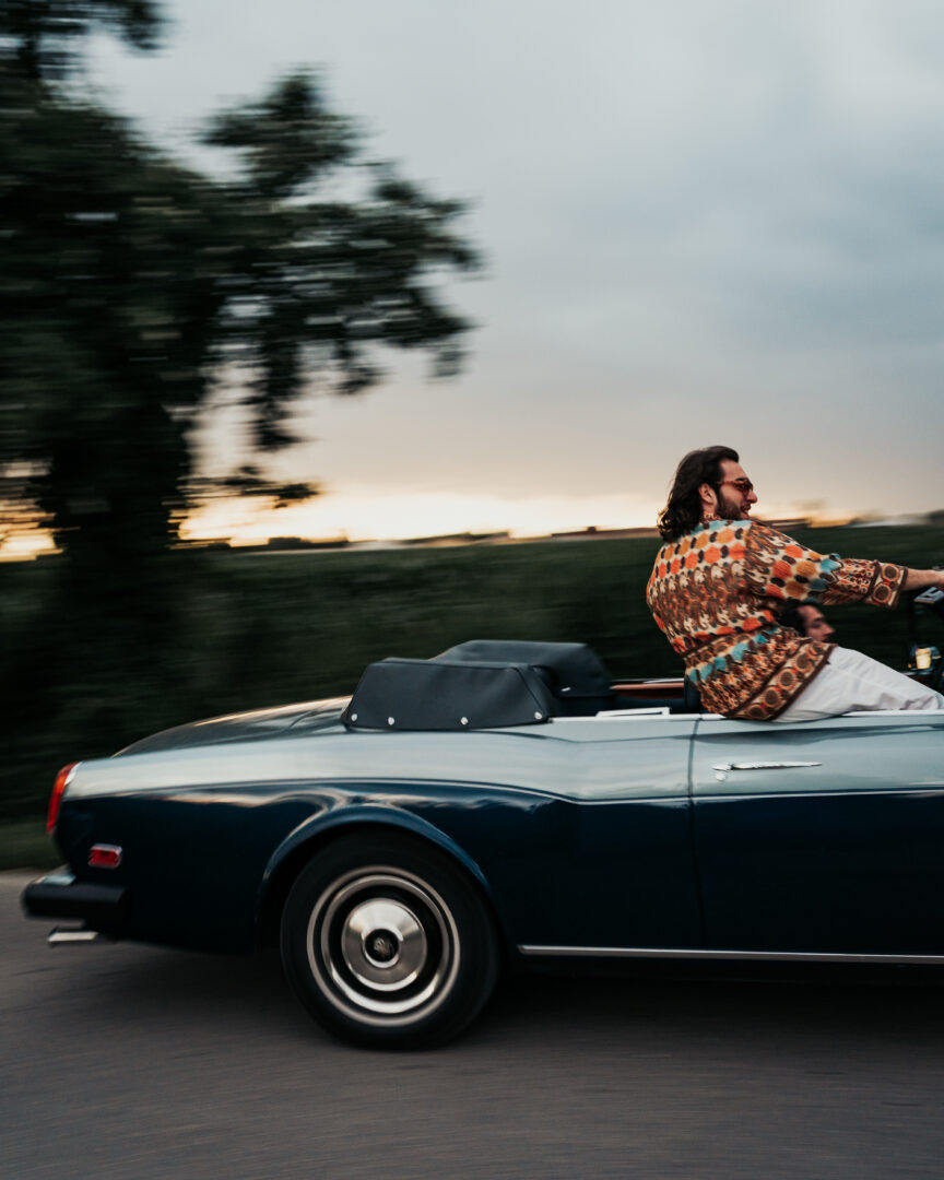 A man wearing a patterned Pantamolle shirt in a classic convertible on a country road at sunset in Italy