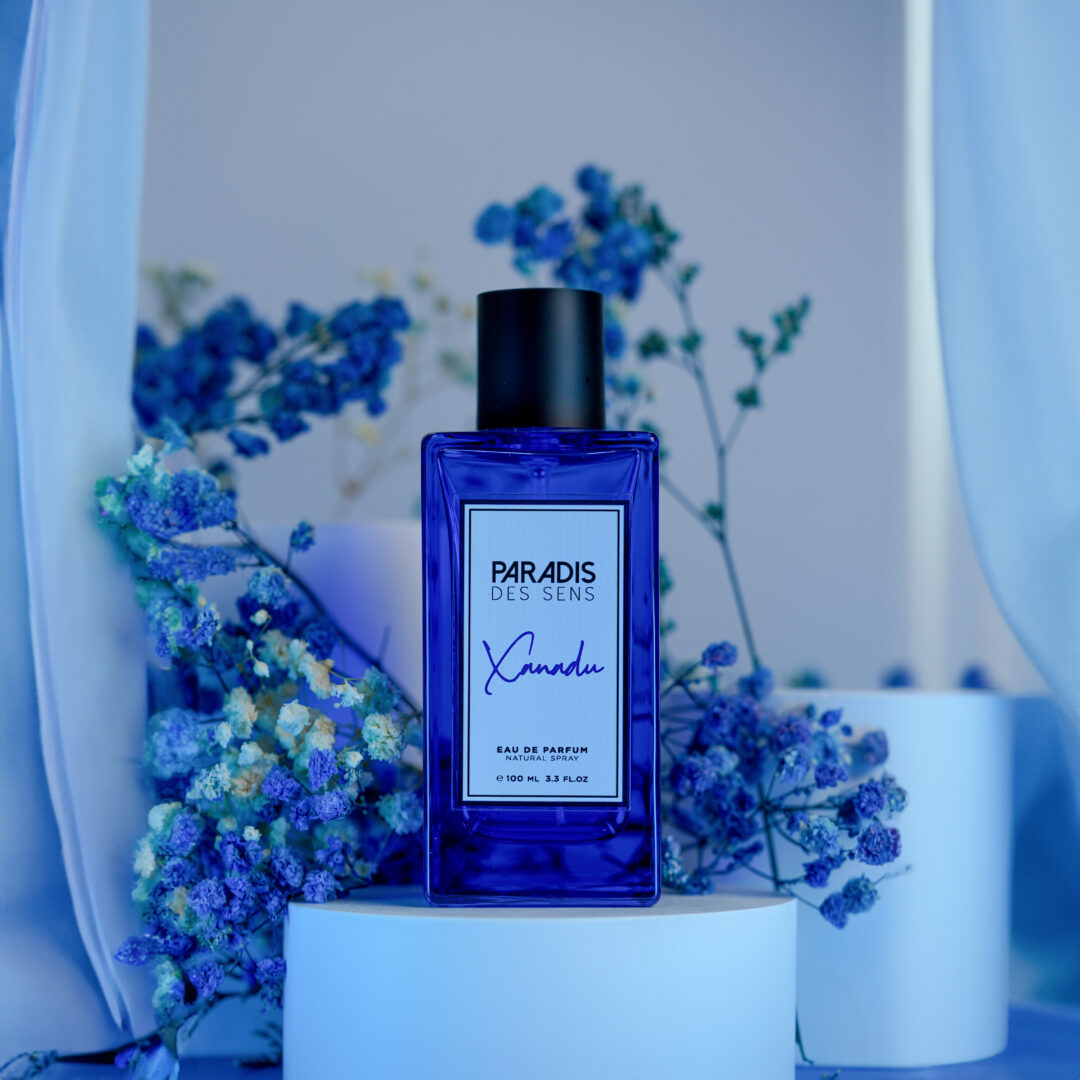 A blue Xanadu bottle by Paradis des Sens on a white pedestal, surrounded by blue flowers.