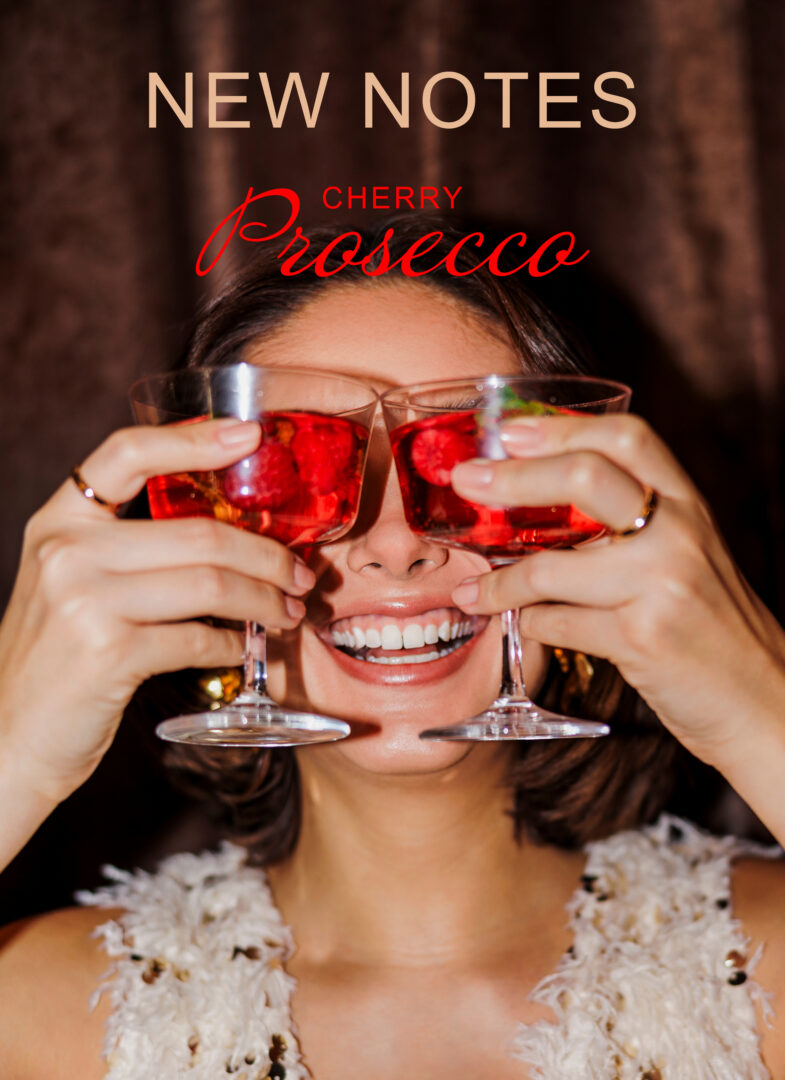 Werbemotiv für Cherry Prosecco von New Notes: Eine lachende Frau hält zwei mit leuchtend rotem Getränk gefüllte Gläser vor ihre Augen. Darüber stehen der Markenname New Notes und der Duftname Cherry Prosecco.