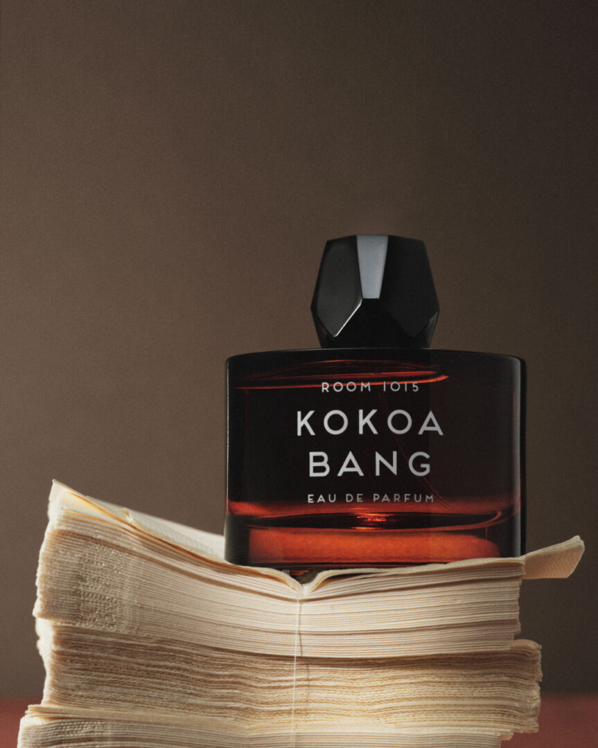 A brown bottle of Room 1015's Kokoa Bang Eau de Parfum with a black cap, placed on an open book against a warm brown background.